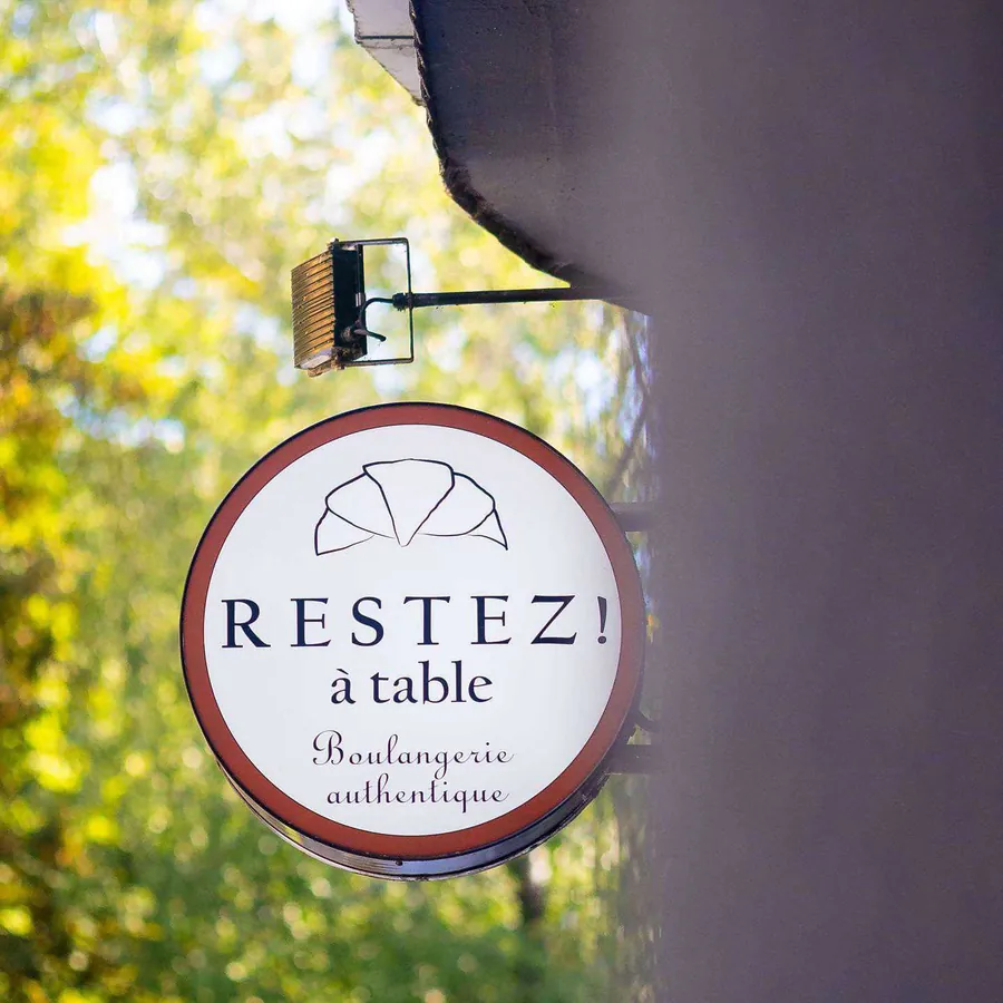 Runder Ausleger „Restez! à table“ an der Hausecke, dahinter grüne Bäume