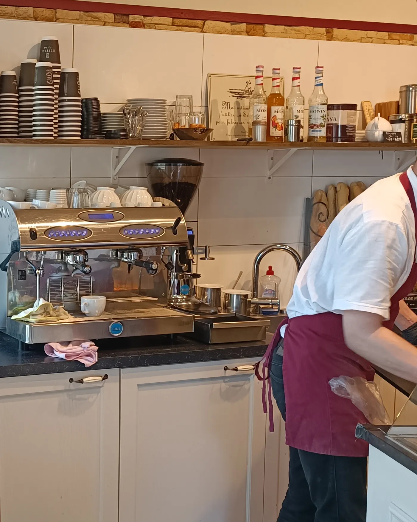 Mitarbeiterin an der Kaffeemaschine hinter der Theke, vorne der Holztresen mit der Getränketafel