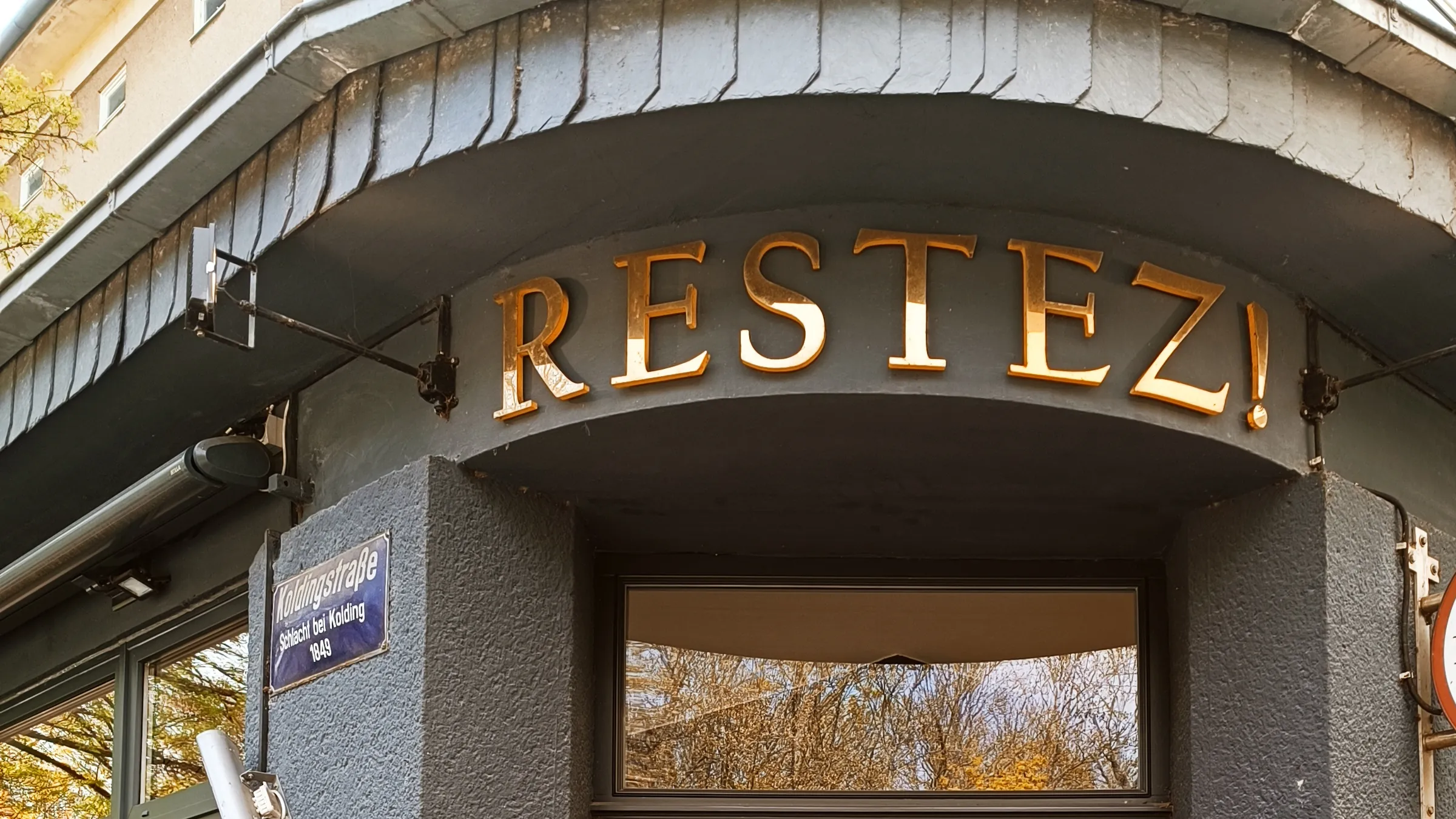 Eingang des Ladens in der Koldingstraße mit dem goldenen Schriftzug „Restez!“, rechts der runde Ausleger „à table“